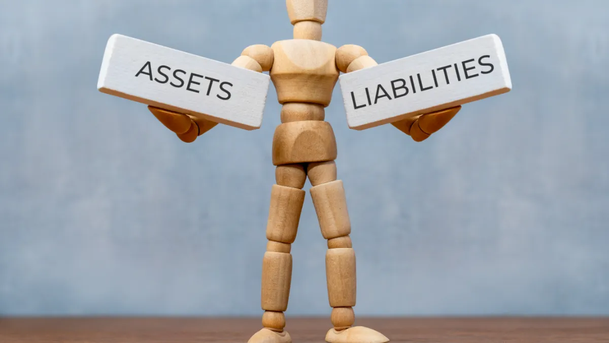 Wooden mannequin holds signs labelled “ASSETS” and “LIABILITIES”.