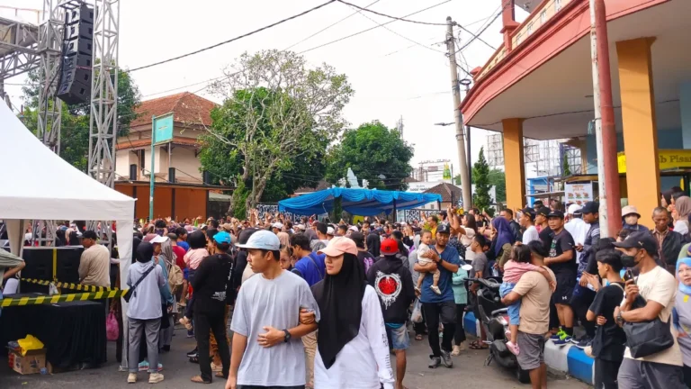 Crowd gathers at an outdoor event in Indonesia, with a stage canopy, speakers and motorcycles lining the street.