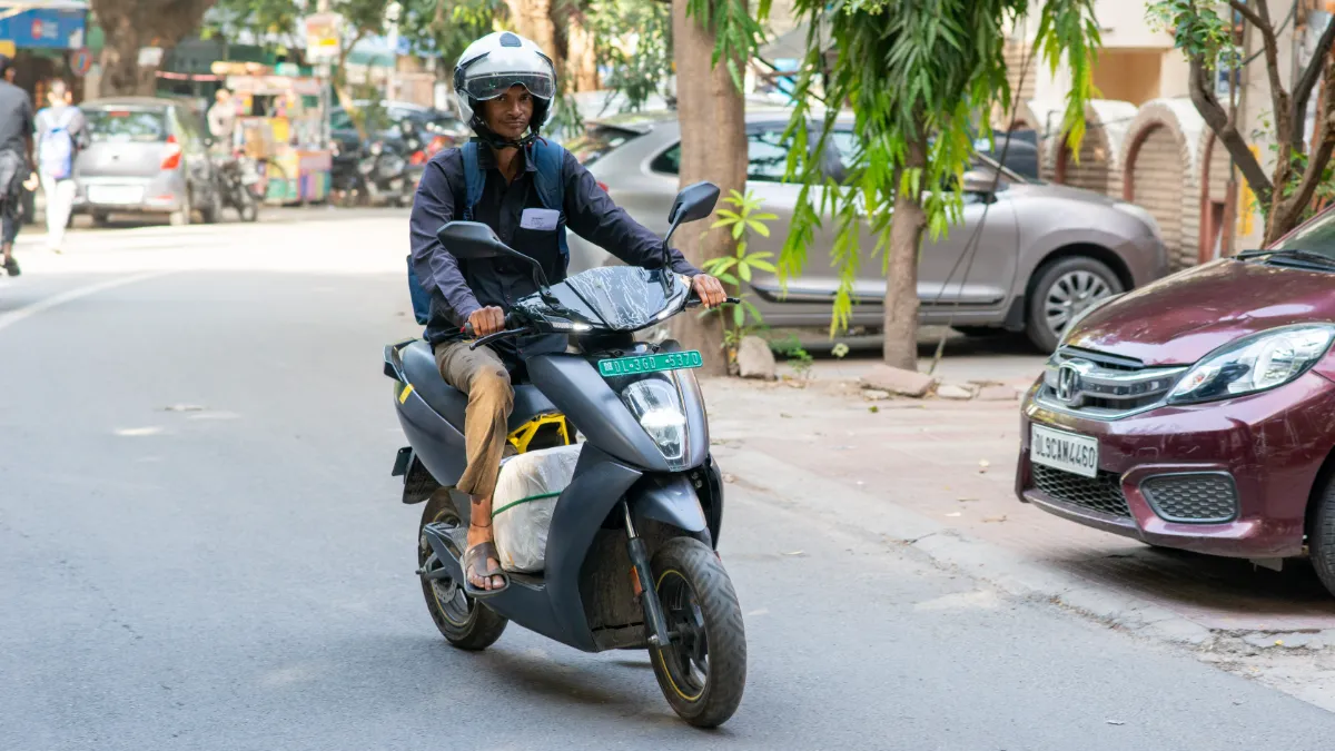 A scooter rider travels along a city street past parked cars and trees.