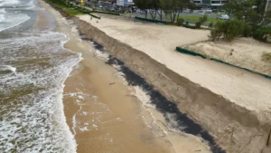 Coastal erosion cuts into a sandy beach beside a road and beachfront buildings.