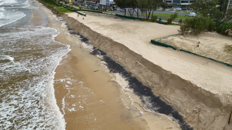 Coastal erosion cuts into a sandy beach beside a road and beachfront buildings.