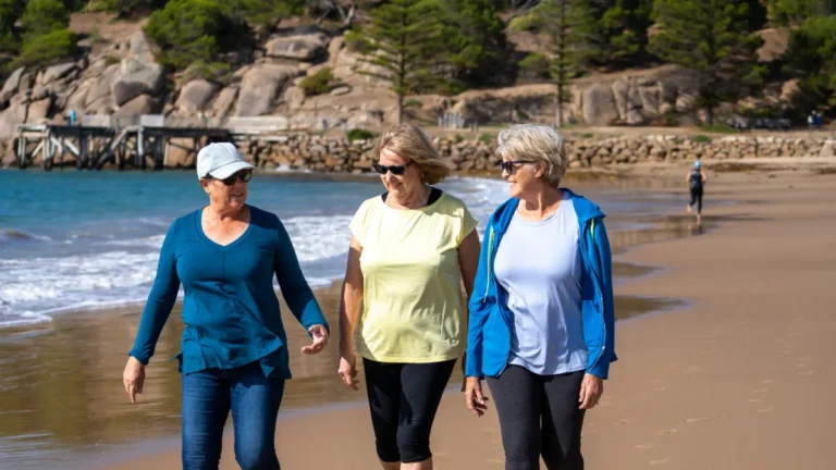 Three older women walk along a beach near the water.