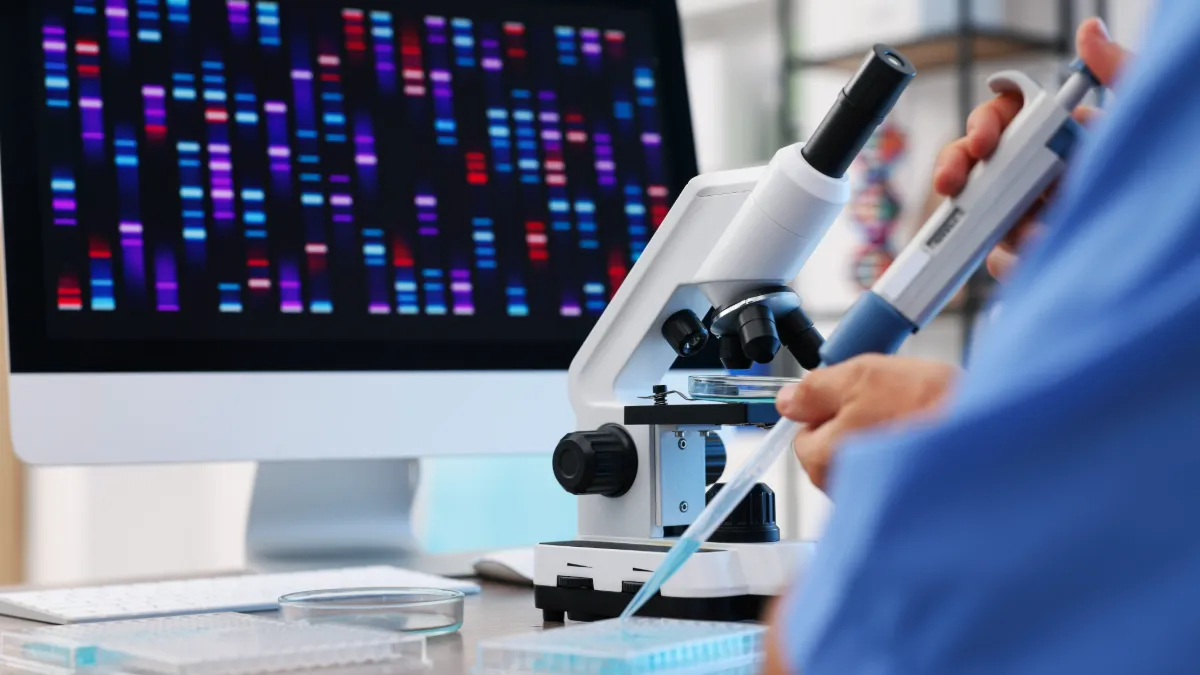 A lab worker examines a sample under a microscope beside a screen displaying colorful genetic sequences.