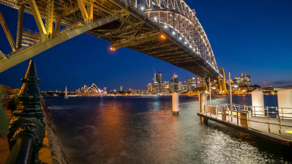 Sydney Harbour Bridge spans the foreground above the harbour, with the Opera House and city skyline illuminated at night.