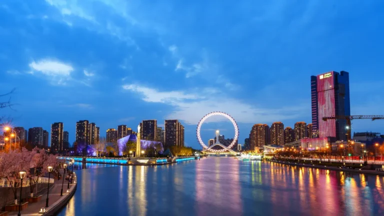 A city skyline and illuminated Ferris wheel overlook a river at dusk.