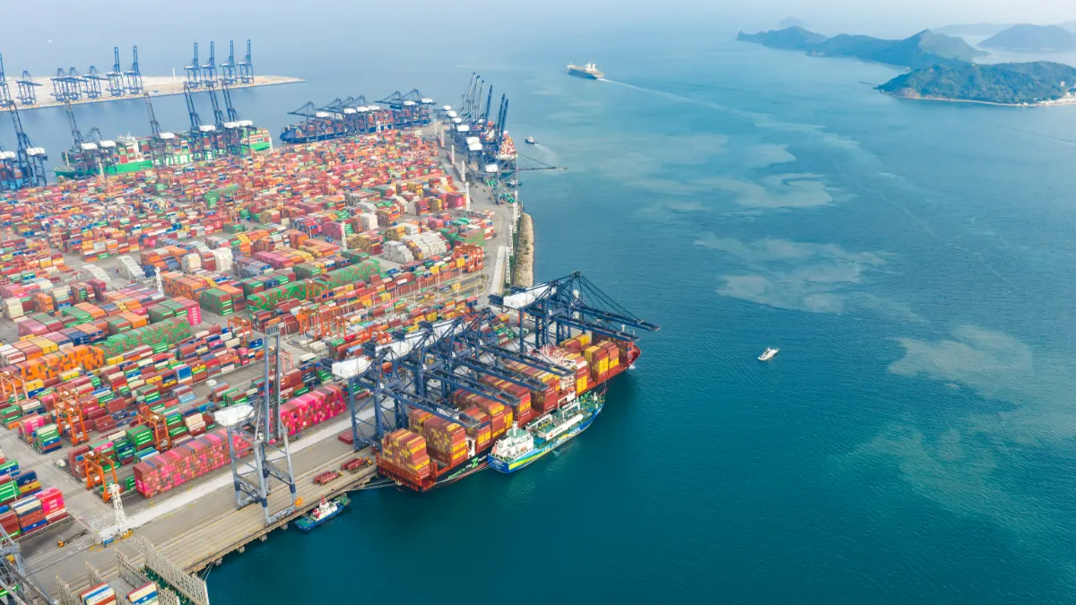 Aerial view of a container port with cranes, stacked cargo containers, ships and islands beyond.