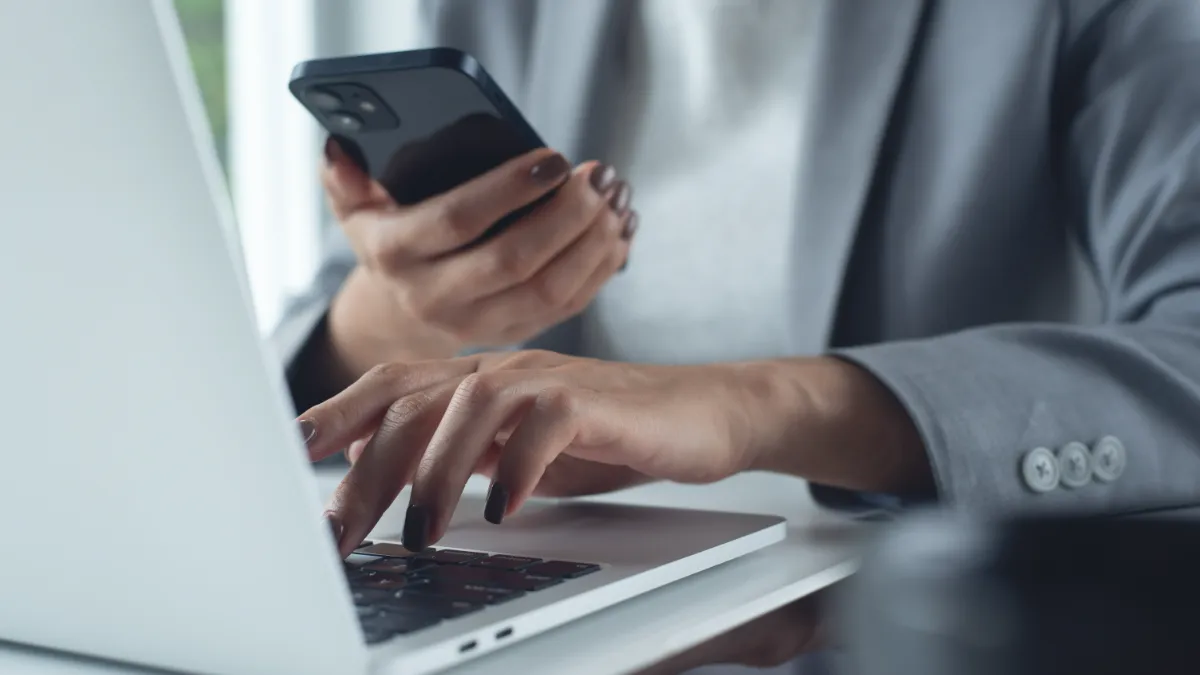Businessperson uses a laptop while holding a smartphone.
