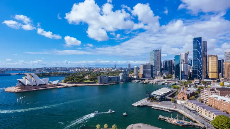 Sydney Harbour with the Opera House and high-rise skyline under a bright, cloud-filled sky.