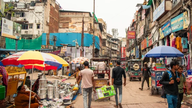 Busy Indian market street with pedestrians, vendors, umbrellas, shops and auto-rickshaws.