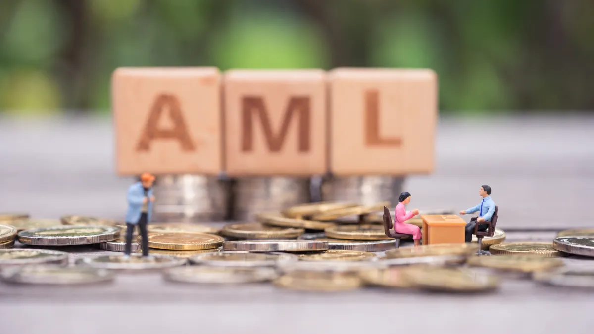 Miniature businesspeople stand and meet among coins beneath wooden blocks spelling “AML.