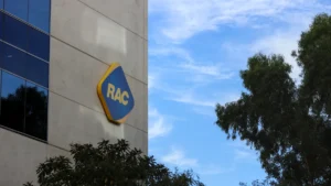 RAC logo mounted on a building exterior beneath a blue sky and beside trees.