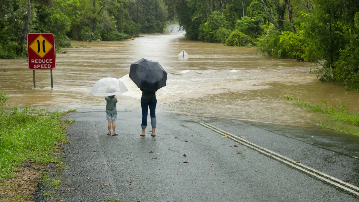 An adult and child stand with umbrellas beside a flooded road and a “Reduce Speed” warning sign.