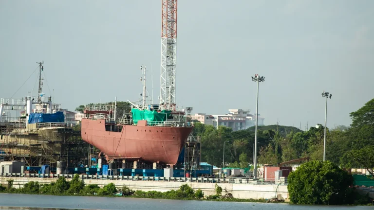 A ship under construction at a waterfront shipyard, surrounded by scaffolding, cranes and trees.
