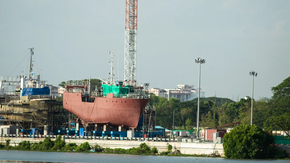 A ship under construction at a waterfront shipyard, surrounded by scaffolding, cranes and trees.