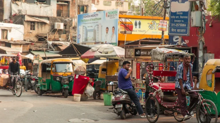 Busy Indian street with auto-rickshaws, a cycle rickshaw, scooters, pedestrians and market signs.