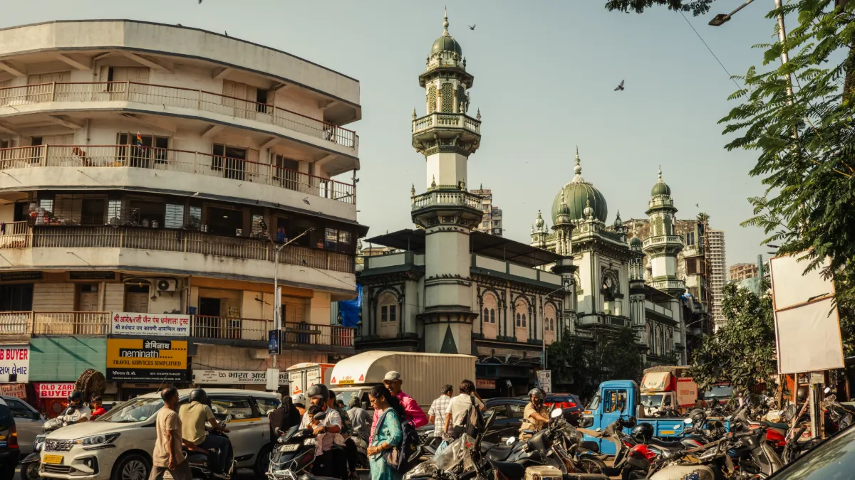 Busy Indian street with motorcycles, cars and pedestrians outside an ornate mosque with domes and minarets.