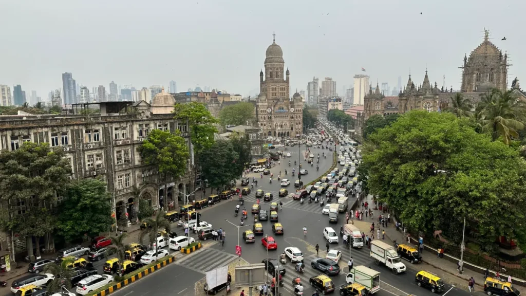 Busy Mumbai intersection with historic Gothic buildings, dense traffic, trees and high-rises in the background.