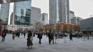 Pedestrians cross a modern urban plaza lined with high-rise office buildings and autumn trees.