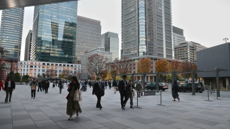 Pedestrians cross a modern urban plaza lined with high-rise office buildings and autumn trees.