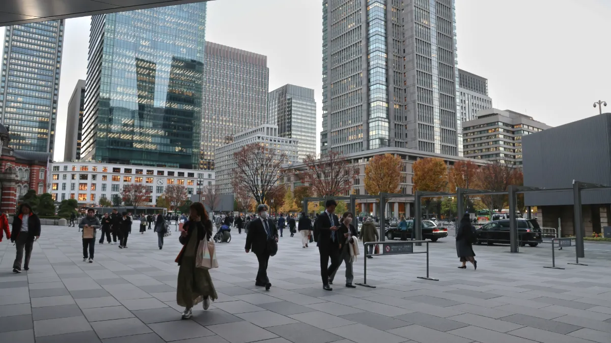 Pedestrians cross a modern urban plaza lined with high-rise office buildings and autumn trees.