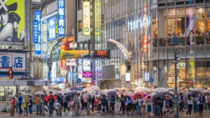 Japan regulator set to suspend new sales at Prudential Life — report Crowds carrying umbrellas gather at a busy Japanese city intersection lined with brightly lit shops and signs.