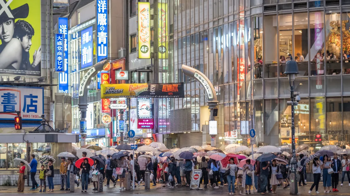 Crowds carrying umbrellas gather at a busy Japanese city intersection lined with brightly lit shops and signs.