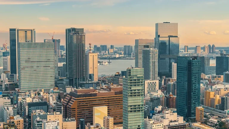 A dense city skyline of high-rise buildings surrounds a waterfront under a hazy sky.