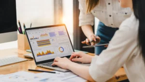 Two businesswomen review data charts on a laptop during a meeting.