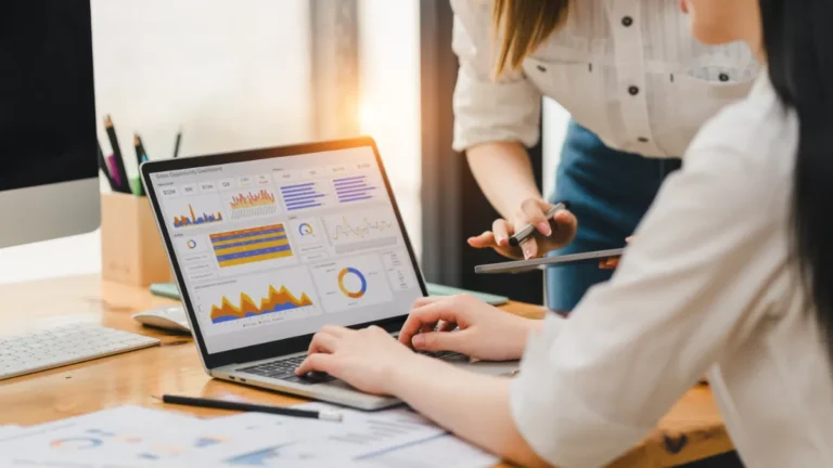 Two businesswomen review data charts on a laptop during a meeting.