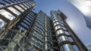 Lloyd’s sets framework for broker-originated reinsurance structures from 2027 Upward view of modern office towers with exposed pipes, metal cladding and curved glass structures.