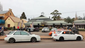 Cars and motorcycles line a street outside The Marketplace as people gather on the footpath.