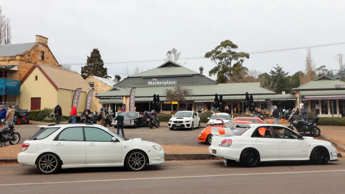 Cars and motorcycles line a street outside The Marketplace as people gather on the footpath.