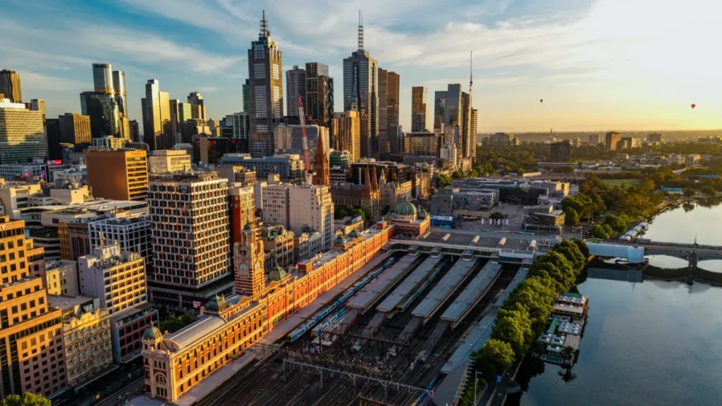 A city skyline with high-rise buildings, a historic station and rail tracks beside a river at sunset.