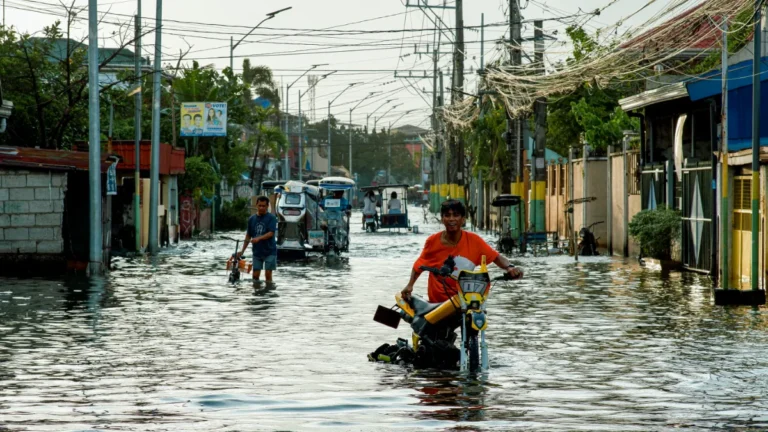People wade through a flooded street as motorized tricycles and a motorcycle navigate the water.