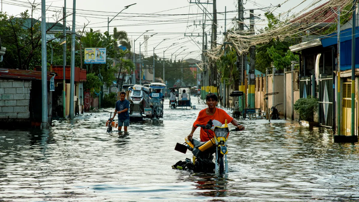 People wade through a flooded street as motorized tricycles and a motorcycle navigate the water.