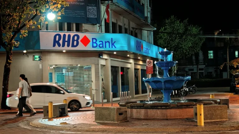 RHB Bank branch with a blue fountain outside at night and pedestrians passing by.