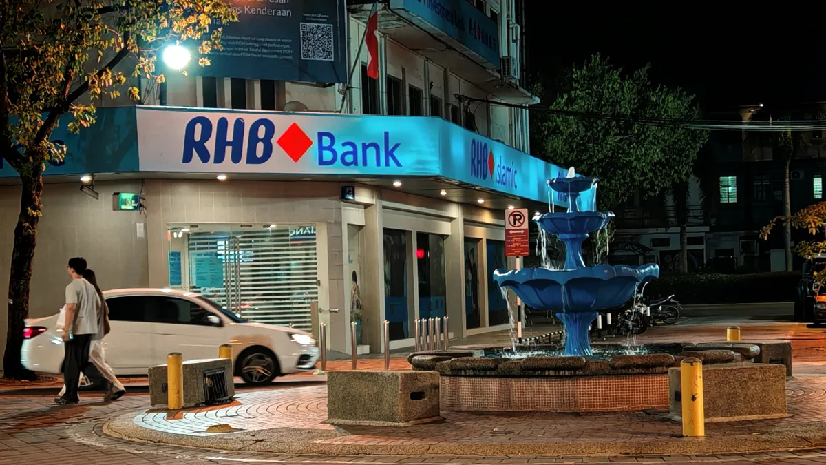 RHB Bank branch with a blue fountain outside at night and pedestrians passing by.