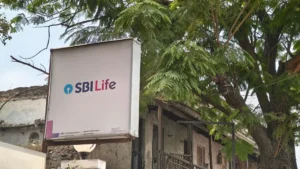 SBI Life Insurance logo on a signboard outside a weathered building beneath leafy trees.