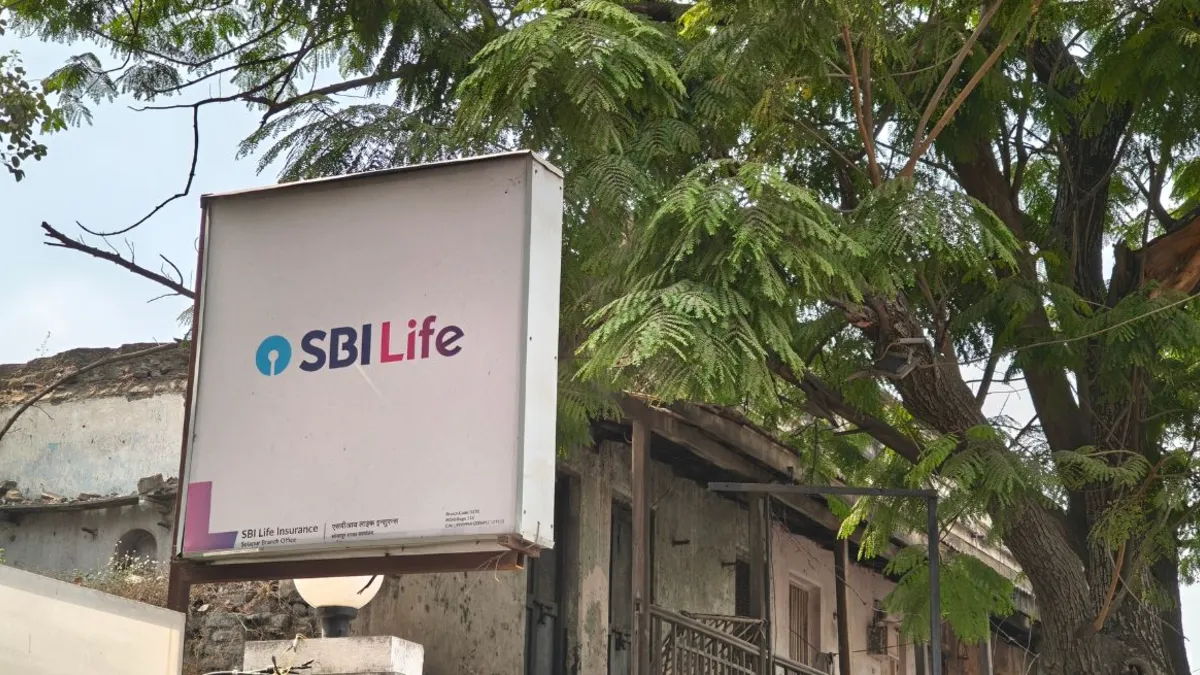 SBI Life Insurance logo on a signboard outside a weathered building beneath leafy trees.