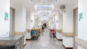 Hospital corridor lined with doors, medical equipment carts and overhead lighting.