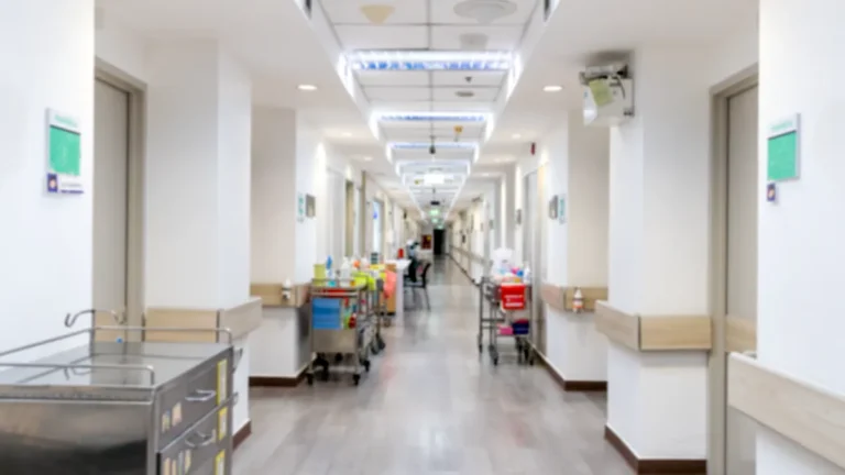 Hospital corridor lined with doors, medical equipment carts and overhead lighting.