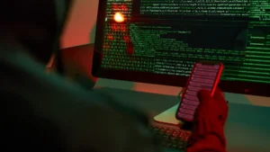 Small and non-US entities face expanding threats as cyber cover gaps persist: Munich Re Person holding a smartphone in front of computer monitors displaying lines of code.