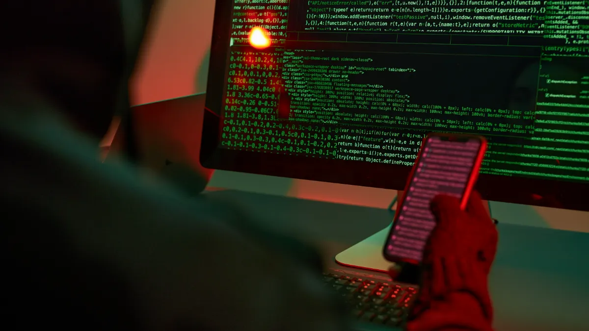 Person holding a smartphone in front of computer monitors displaying lines of code.