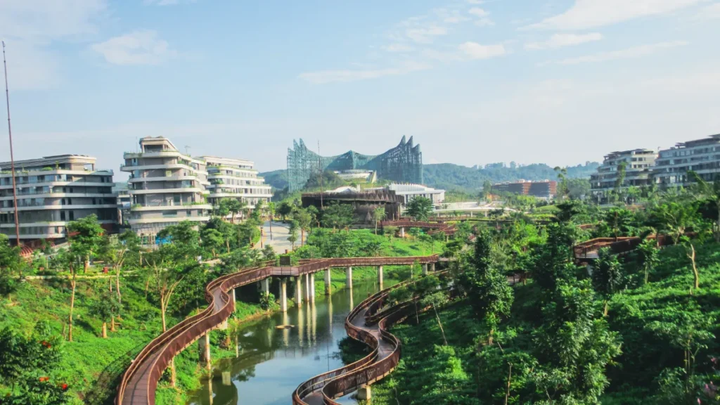 Modern buildings surround a lush park with winding wooden walkways over a narrow waterway.