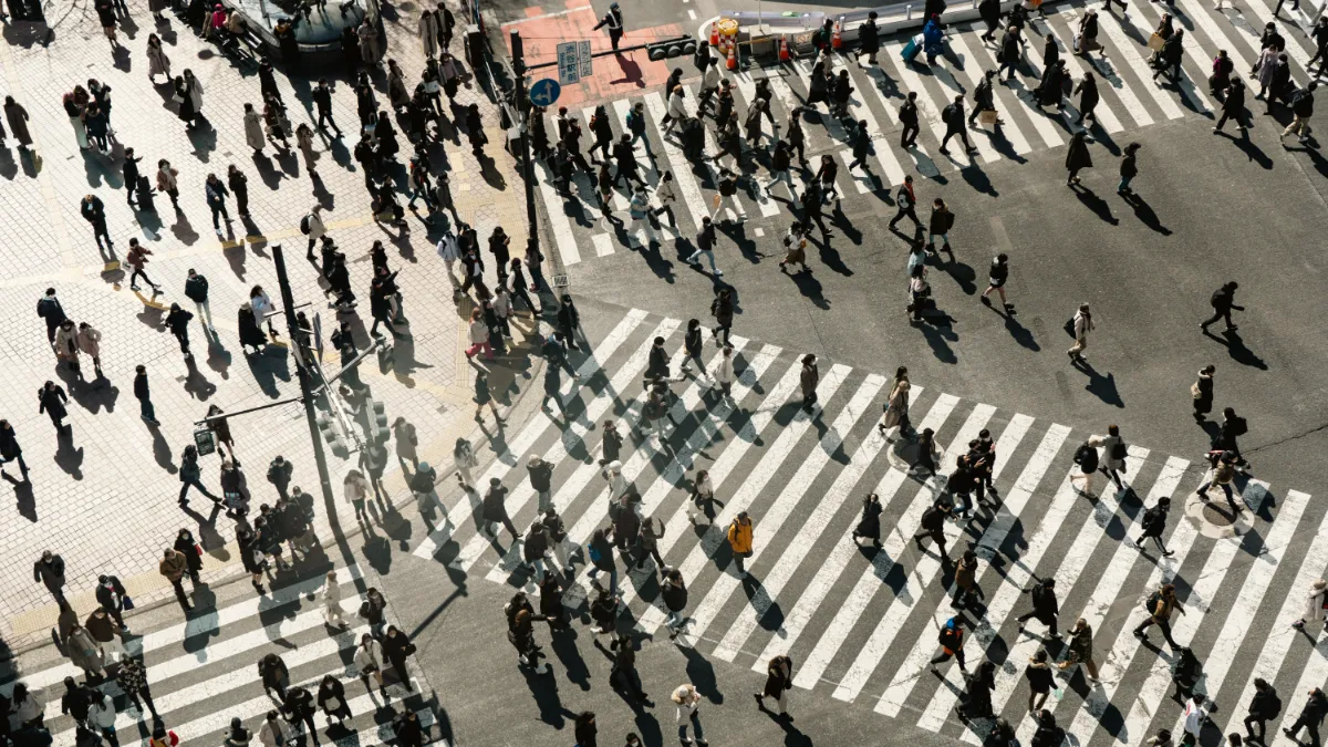 Crowds of pedestrians cross a wide city intersection in bright sunlight, casting long shadows.