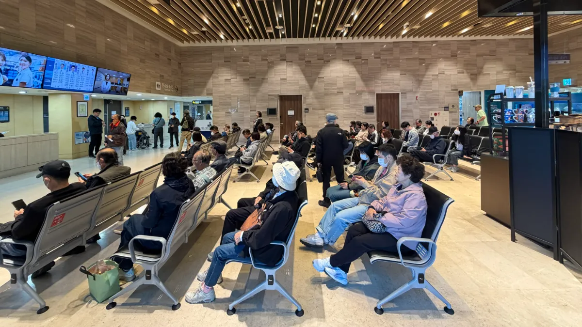 Crowded medical waiting room with people seated in rows beneath digital screens.