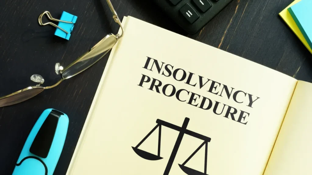 Open book titled “Insolvency Procedure” beside glasses, binder clips and a calculator on a desk.