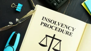 Open book titled “Insolvency Procedure” beside glasses, binder clips and a calculator on a desk.