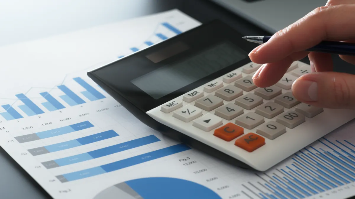 A hand uses a calculator over printed financial charts and graphs.