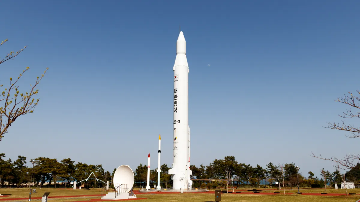 A large white rocket stands on display outdoors beside two smaller rockets under a clear blue sky.